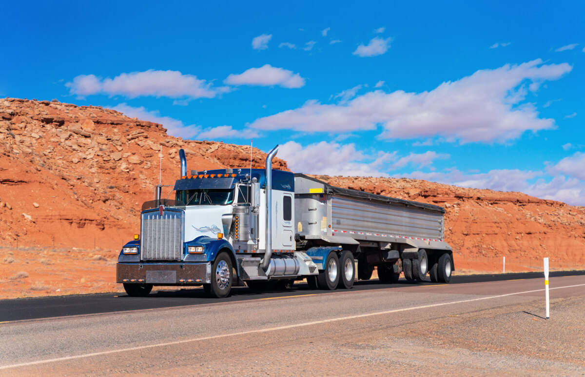 White and blue dump truck on a rural road next to a pile of fill dirt, to show insurance coverage for dump truck.